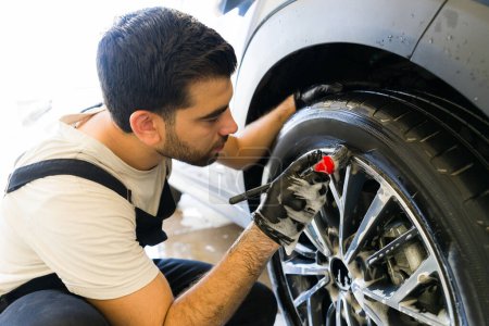 Foto de Joven hombre brillando los neumáticos con un cepillo mientras se lava el coche en el servicio de detalle automático - Imagen libre de derechos