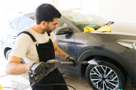 Foto de Hombre latino joven limpiando el coche con una lavadora de alta presión en el lavado de coches - Imagen libre de derechos