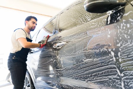 Foto de Hombre mexicano de 20 años que trabaja en el lavado de autos lavando un coche cliente con jabón y guante de microfibra - Imagen libre de derechos