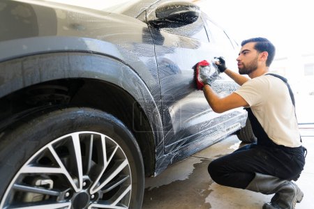 Téléchargez les photos : Hispanique mâle travaillant au lavage de voiture laver une voiture de client avec du savon et un gant en microfibre - en image libre de droit