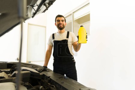 Téléchargez les photos : Beau jeune homme latin travaillant au garage automobile tenant l'huile moteur peut faire un contact visuel - en image libre de droit