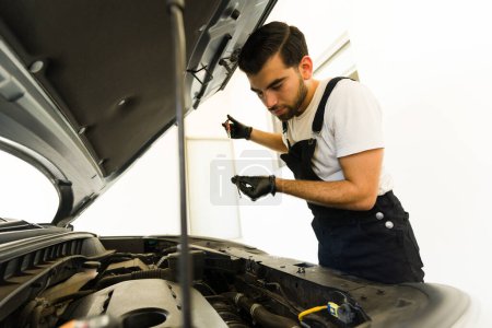 Téléchargez les photos : Jeune mécanicien automobile latin mâle vérifiant le niveau d'huile moteur d'une voiture - en image libre de droit