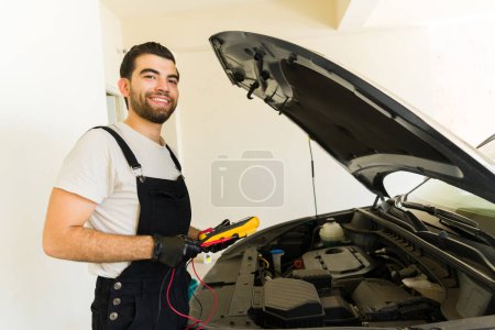 Photo for Young Latin mechanic holding a multimeter to check a battery at an auto shop smiling and making eye contact - Royalty Free Image