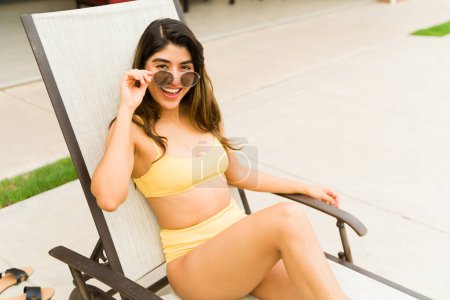 Téléchargez les photos : Femme magnifique excitée dans un bikini portant des lunettes de soleil tout en se relaxant dans la chaise longue de la piscine se sentant heureux - en image libre de droit