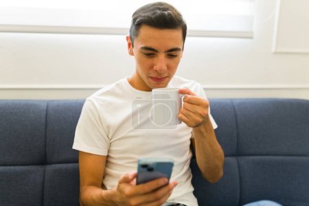 Téléchargez les photos : Homme attrayant buvant du café dans une tasse blanche 11 tout en portant un t-shirt blanc maquillé et textos sur le smartphone à la maison - en image libre de droit