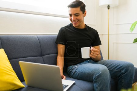 Téléchargez les photos : Joyeux jeune homme travaillant à la maison en utilisant l'ordinateur portable tout en portant un t-shirt maquillé à l'aide d'une tasse de café - en image libre de droit