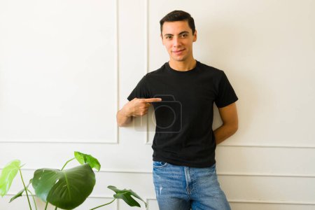 Foto de Hombre joven caucásico sonriendo mientras señala una camiseta negra de maqueta con un diseño de logotipo con espacio para copiar - Imagen libre de derechos