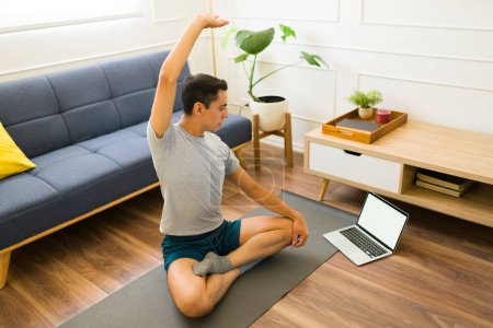 Téléchargez les photos : Jeune homme actif portant un t-shirt gris maquillé étirant tout en faisant du yoga à l'aide de son ordinateur portable avec un écran vide avec espace de copie - en image libre de droit