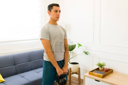 Téléchargez les photos : Jeune homme actif soulevant un poids de kettlebell portant un t-shirt gris maquillé à la maison - en image libre de droit