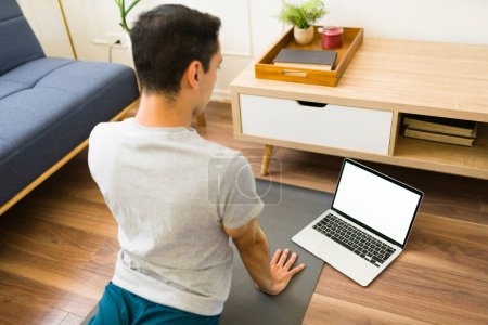 Téléchargez les photos : Vue arrière d'un jeune homme en forme faisant du yoga à la maison en regardant une vidéo sur l'écran blanc de l'ordinateur portable avec espace de copie - en image libre de droit