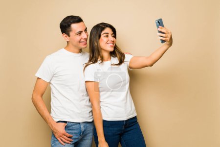 Téléchargez les photos : Couple heureux souriant portant des t-shirts maquettes assortis avec espace de copie prenant un selfie avec un smartphone - en image libre de droit