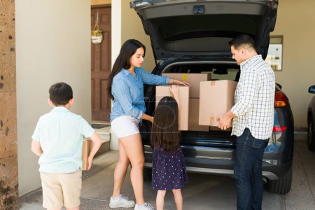 Photo for Latin family getting boxes out of the car trunk to unpack while moving into a new house with the help of their children - Royalty Free Image