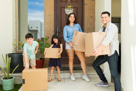 Photo for Beautiful happy family with children smiling while carrying boxes in the entryway of their new big house while moving in together - Royalty Free Image