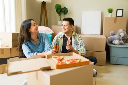 Foto de Atractiva pareja feliz riendo juntos mientras comen pizza para la cena en cajas de cartón después de desempacar en su nueva casa o apartmnet - Imagen libre de derechos