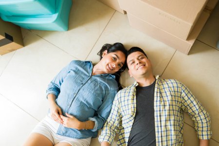 Téléchargez les photos : Vue de dessus d'un couple détendu reposant sur le sol et prenant une pause des boîtes de déballage et des meubles mobiles dans leur nouvelle maison - en image libre de droit