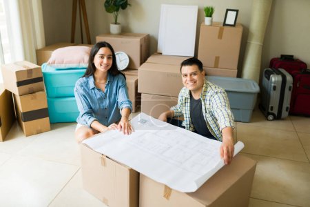 Téléchargez les photos : Heureux beau couple souriant tout en regardant les plans de la maison après avoir emménagé dans leur nouvelle maison avec un crédit hypothécaire - en image libre de droit