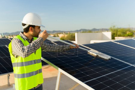 Foto de Ingeniero latino con chaleco y casco limpiando los paneles solares en la azotea para generar energía limpia - Imagen libre de derechos