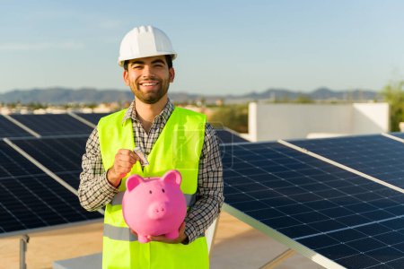Foto de Retrato de un feliz ingeniero latino usando una alcancía mientras ahorra energía con la instalación del panel solar - Imagen libre de derechos