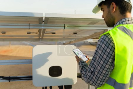 Téléchargez les photos : Ingénieur professionnel vérifiant le moniteur d'énergie sur le smartphone tout en travaillant dans l'installation de panneaux solaires - en image libre de droit
