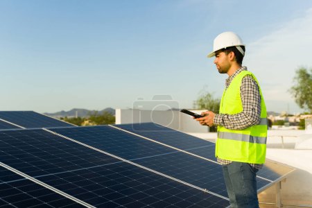 Foto de Ingeniero latino con chaleco verde y casco comprobando la energía en los paneles solares y células fotovoltaicas utilizando una tableta - Imagen libre de derechos