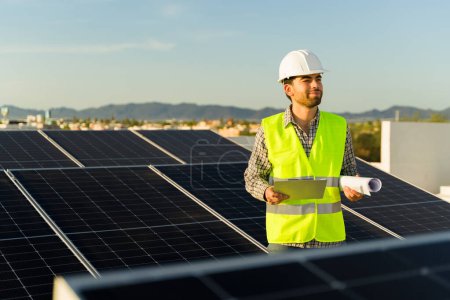Téléchargez les photos : Travailleur ingénieur heureux souriant tout en tenant un presse-papiers et des plans d'installation des panneaux solaires à énergie propre - en image libre de droit