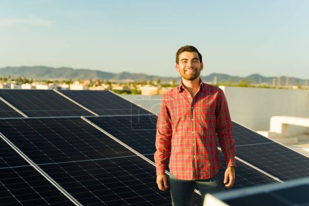 Téléchargez les photos : Latino jeune homme souriant tout en regardant heureux d'utiliser l'énergie propre à la maison avec ses panneaux solaires et cellules photovoltaïques - en image libre de droit