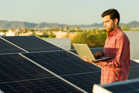 Foto de Hombre latino guapo con un portátil usando energía limpia en casa y comprobando sus paneles solares en la azotea - Imagen libre de derechos