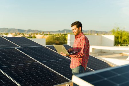 Foto de Feliz hombre hispano revisando el monitor de energía en el portátil mientras usa paneles solares y células fotovoltaicas en casa - Imagen libre de derechos