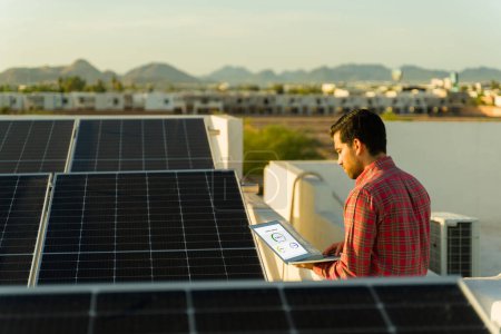 Photo for Latin young man seen from behind using the laptop to check the energy monitor of his residential solar panels in the rooftop - Royalty Free Image