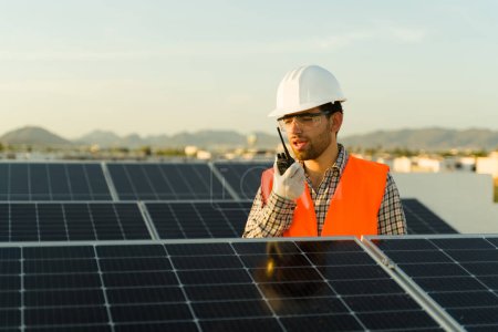 Foto de Ingeniero hombre atractivo con un chaleco naranja y casco hablando en el walkie talkie sobre la instalación de paneles solares y células fotovoltaicas - Imagen libre de derechos