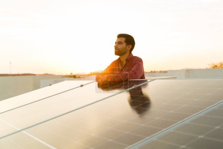 Foto de Hombre latino relajado y dueño de casa en la azotea mirando feliz sobre el uso de paneles solares y energía sostenible limpia - Imagen libre de derechos