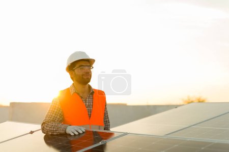 Foto de Guapo ingeniero feliz hombre con gafas de seguridad sonriendo mientras trabaja en la azotea con paneles solares y células fotovoltaicas - Imagen libre de derechos