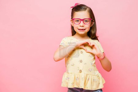 Foto de Niña cariñosa con gafas haciendo un corazón y dando amor a su familia y amigos contra el espacio de copia de fondo rosa - Imagen libre de derechos
