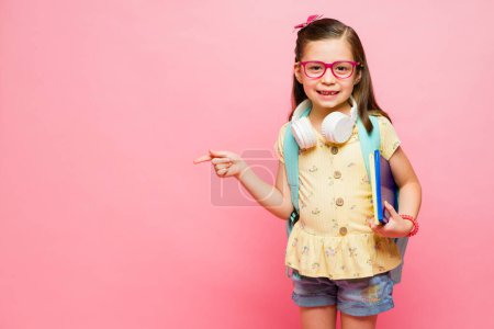 Téléchargez les photos : Petite fille intelligente apprenant à l'école portant son sac à dos et souriant tout en pointant vers un espace de copie rose - en image libre de droit