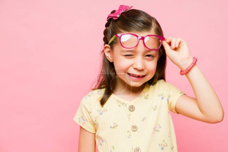 Foto de Alegre niña sonriendo mientras se quita las gafas y guiño mirando feliz divertirse contra un fondo rosa - Imagen libre de derechos