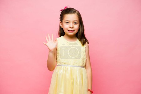 Téléchargez les photos : Joyeux caucasien petite fille avec une jolie robe jaune souriant bonjour ou au revoir à ses amis regardant heureux - en image libre de droit