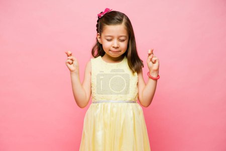 Téléchargez les photos : Adorable petite fille de 7 ans se sentant chanceuse en croisant les doigts et en demandant que ses rêves se réalisent et en pensant à ses souhaits - en image libre de droit