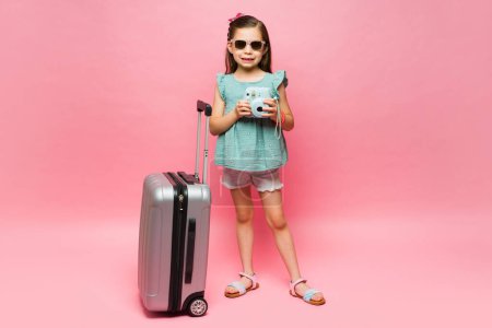 Téléchargez les photos : Beau petit enfant avec des lunettes de soleil à l'aide d'un appareil photo instantané et valise prête à partir en vacances d'été - en image libre de droit
