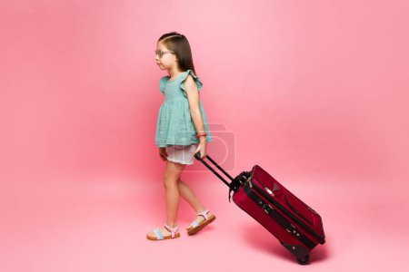 Photo for Adorable 7 year old little girl going on summer vacations walking with her suitcase in front of a pink copy space background - Royalty Free Image