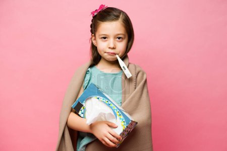 Téléchargez les photos : Malade enfant caucasien de 7 ans souffrant de fièvre ou enroulé à froid dans une couverture et utilisant un thermomètre et une boîte de tissus - en image libre de droit