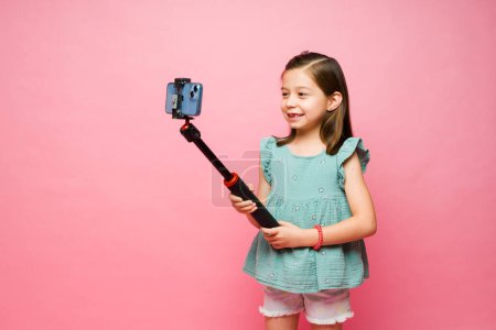 Photo for Adorable cute little girl smiling while using a smartphone filming a video with a selfie stick or taking pictures - Royalty Free Image