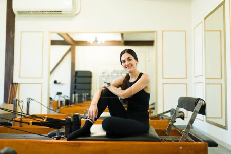 Téléchargez les photos : Joyeux belle femme souriant tout prêt pour sa classe de lit réformateur pilates dans une belle salle de gym haut de gamme - en image libre de droit