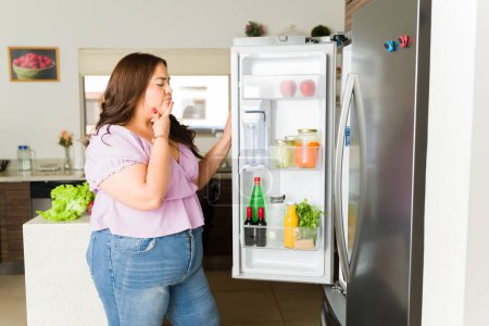 Téléchargez les photos : Une jeune femme latine réfléchie réfléchissant à ce qu'il faut manger pour le déjeuner et regardant le réfrigérateur ouvert dans la cuisine à la recherche coûteuse - en image libre de droit
