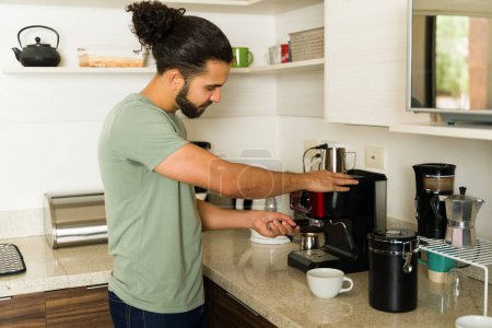 Photo for Handsome young man smiling while preparing coffee in the kitchen and getting ready for house chores at home - Royalty Free Image