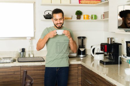 Téléchargez les photos : Joyeux jeune homme souriant tout en appréciant de boire une tasse de café dans la cuisine et se préparer à faire des travaux ménagers à la maison - en image libre de droit