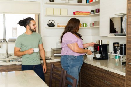 Photo for Latin woman using the coffee maker preparing coffee in the kitchen with her smiling boyfriend enjoying domestic life together - Royalty Free Image