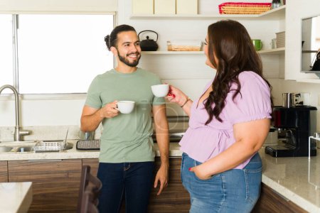 Téléchargez les photos : Heureux jeune homme et femme souriant regardant gai tout en buvant du café ensemble dans la cuisine se préparer à faire des tâches ménagères - en image libre de droit