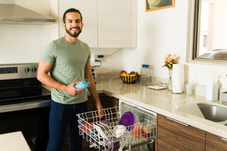 Foto de Atractivo hombre mexicano sonriendo mirando a la cámara cargando el lavavajillas para lavar los platos y hacer las tareas de la casa luciendo feliz - Imagen libre de derechos