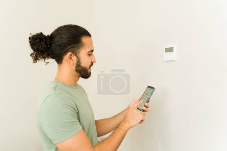 Téléchargez les photos : Heureux jeune homme contrôlant le thermostat ac et le thermostat de climatisation tout en utilisant l'application smartphone à la maison - en image libre de droit
