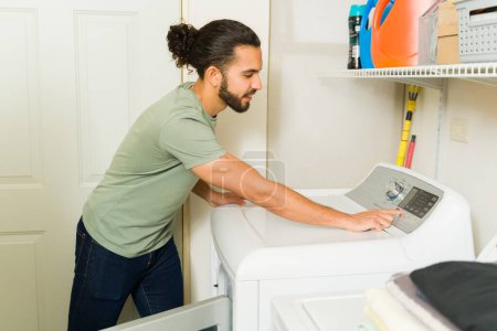 Photo for Attractive hispanic man turning on the washing machine at home and ready to do laundry and house chores - Royalty Free Image